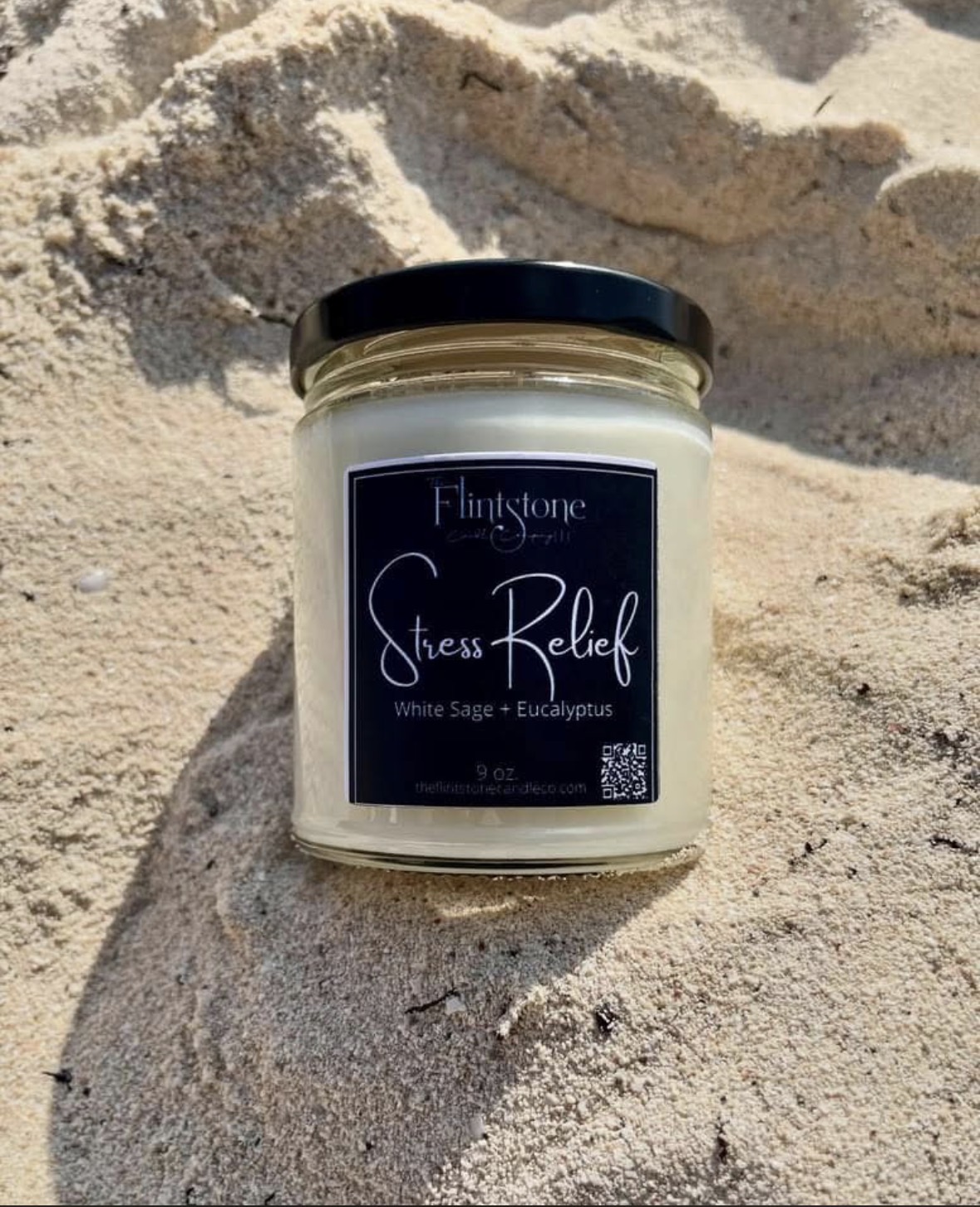 A candle jar labeled 'Stress Relief' with white sage and eucalyptus, placed on sandy ground.