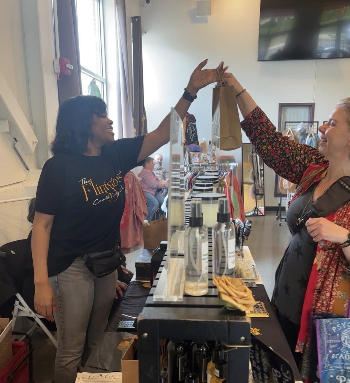 Two women exchange a high-five behind a counter in a bright indoor space, with people and a TV in the background.