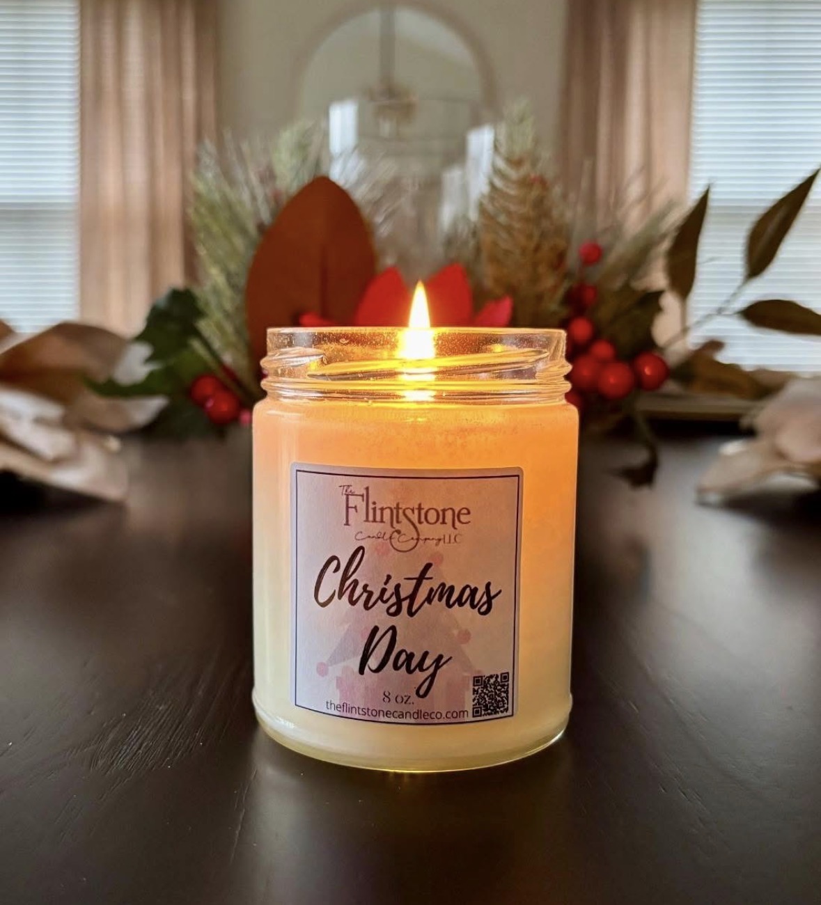 Lit candle in a jar with a Christmas label, in front of holiday decorations and a window, on a dark table.