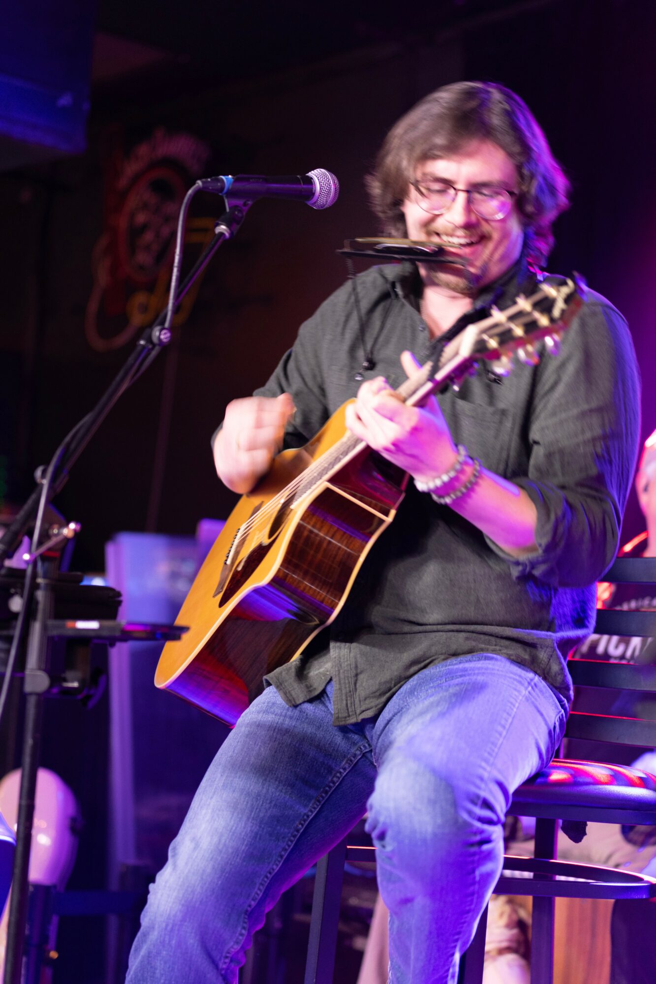 Person playing guitar and holding a harmonica, smiling, on stage with microphone, purple lighting, seated on a stool.