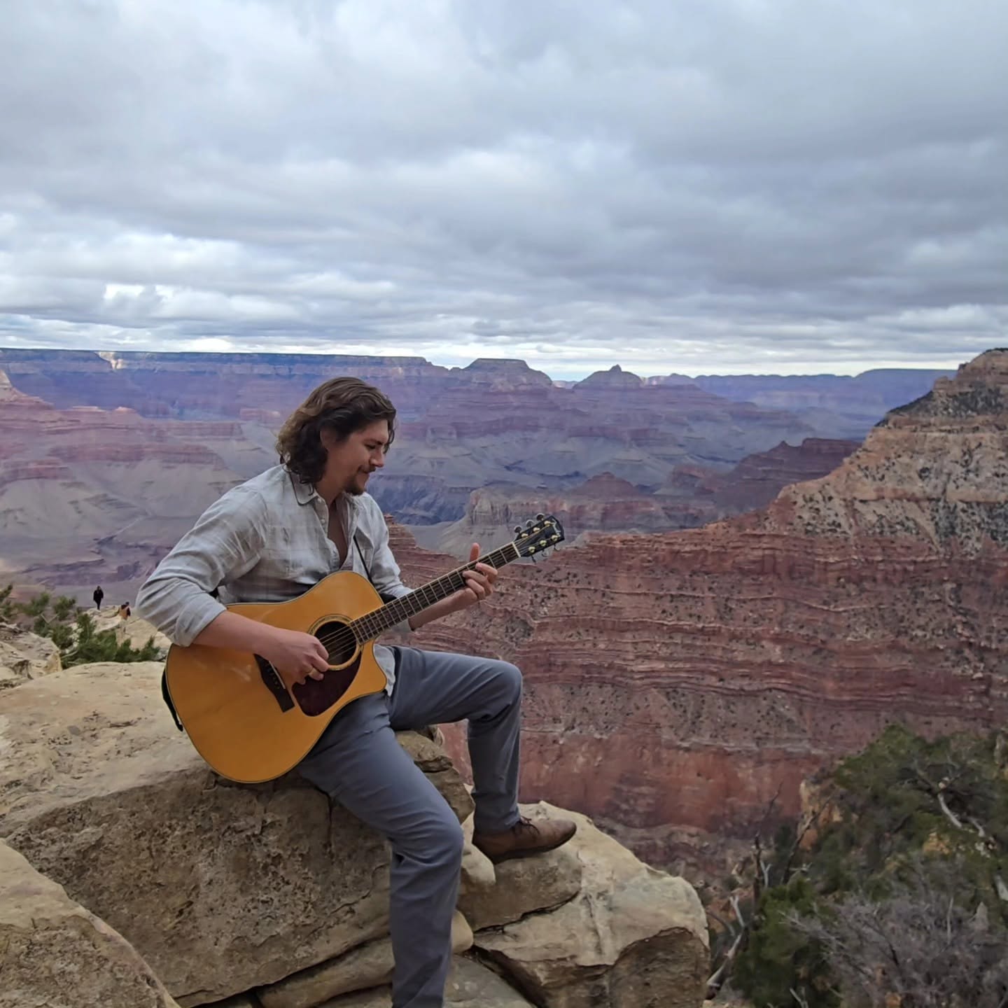 Person sitting on rock playing guitar at Grand Canyon with cloudy sky in background.