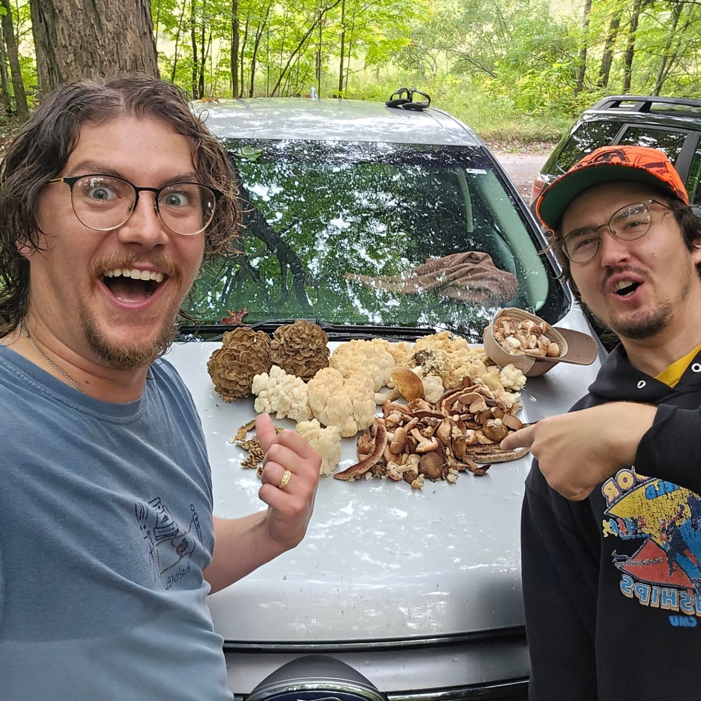 Two men outdoors with a car, smiling, pointing at food on the car's hood, surrounded by trees.
