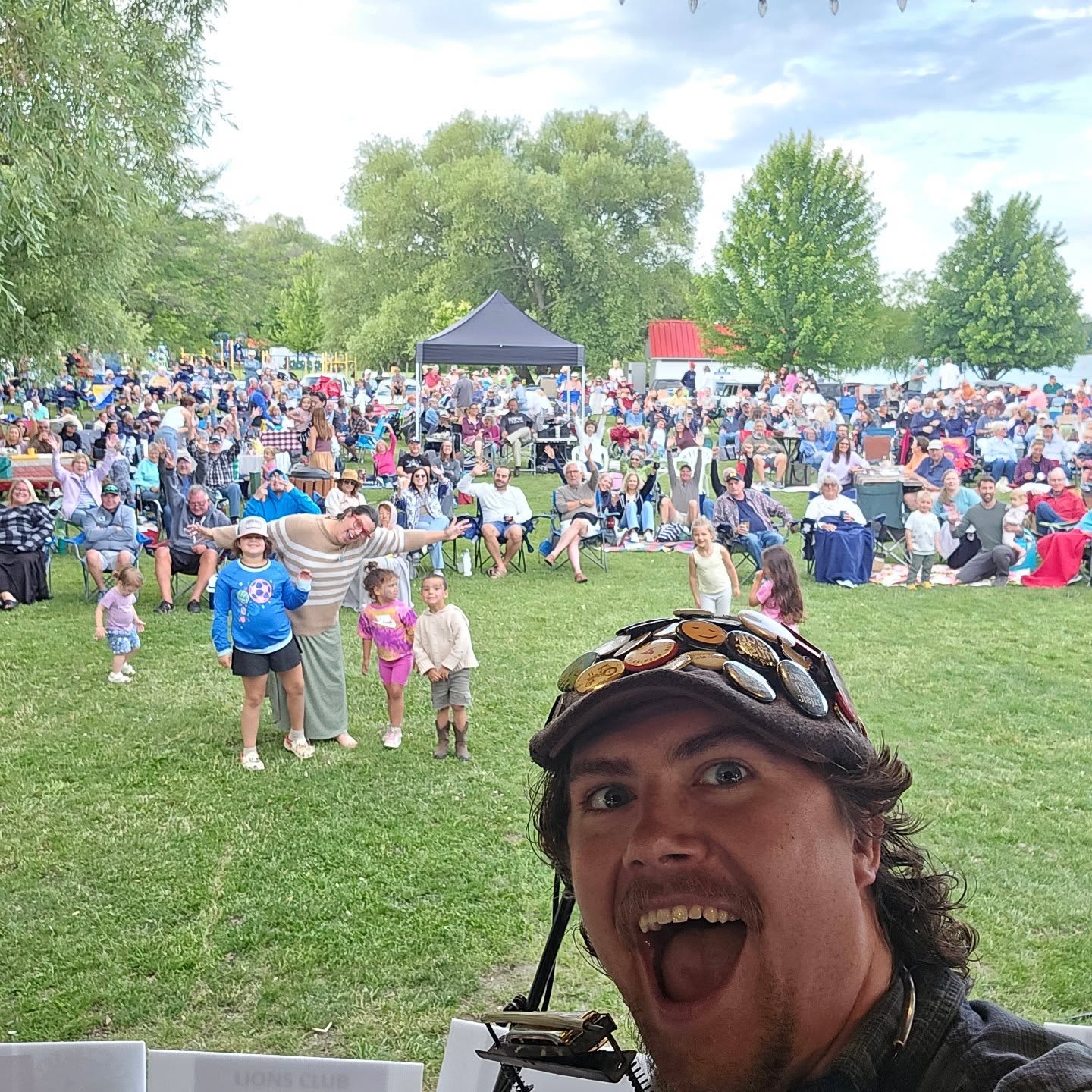 Man taking selfie outdoors with a large crowd and trees in the background, smiling widely.
