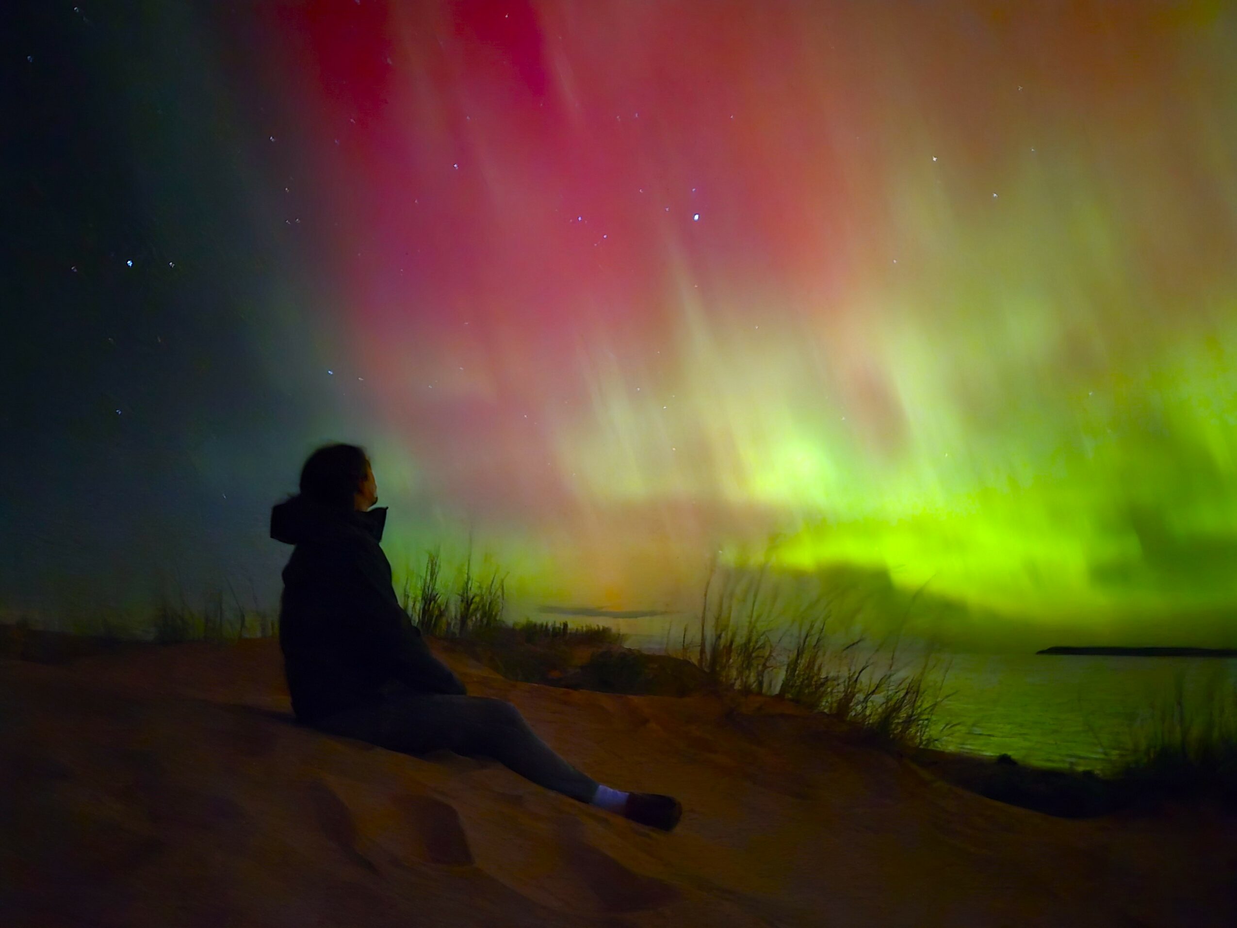 Person sitting on a sand dune watching colorful northern lights in the night sky, with stars visible.