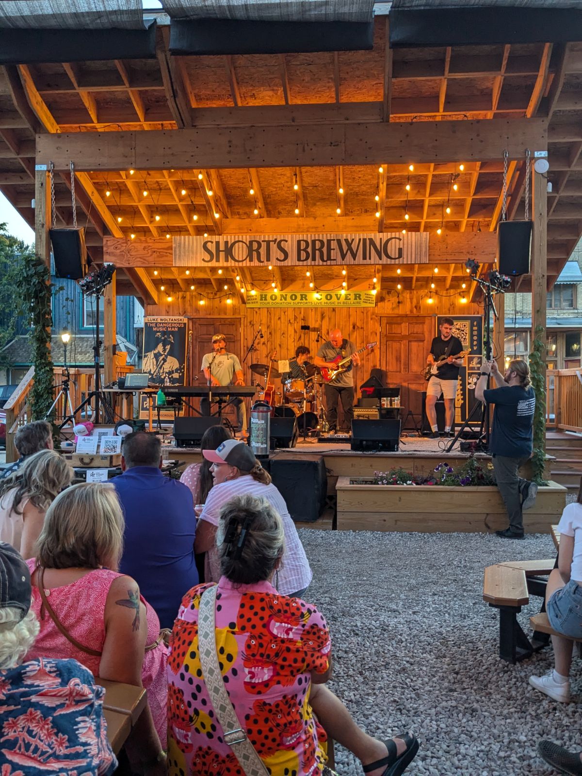 Outdoor stage with a band performing, audience seated on benches, wooden structure with a sign reading 'SHORT'S BREWING', evening setting.