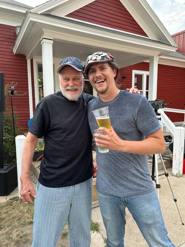 Two men smiling outdoors in front of a red house with white porch columns, one holding a glass of beer.
