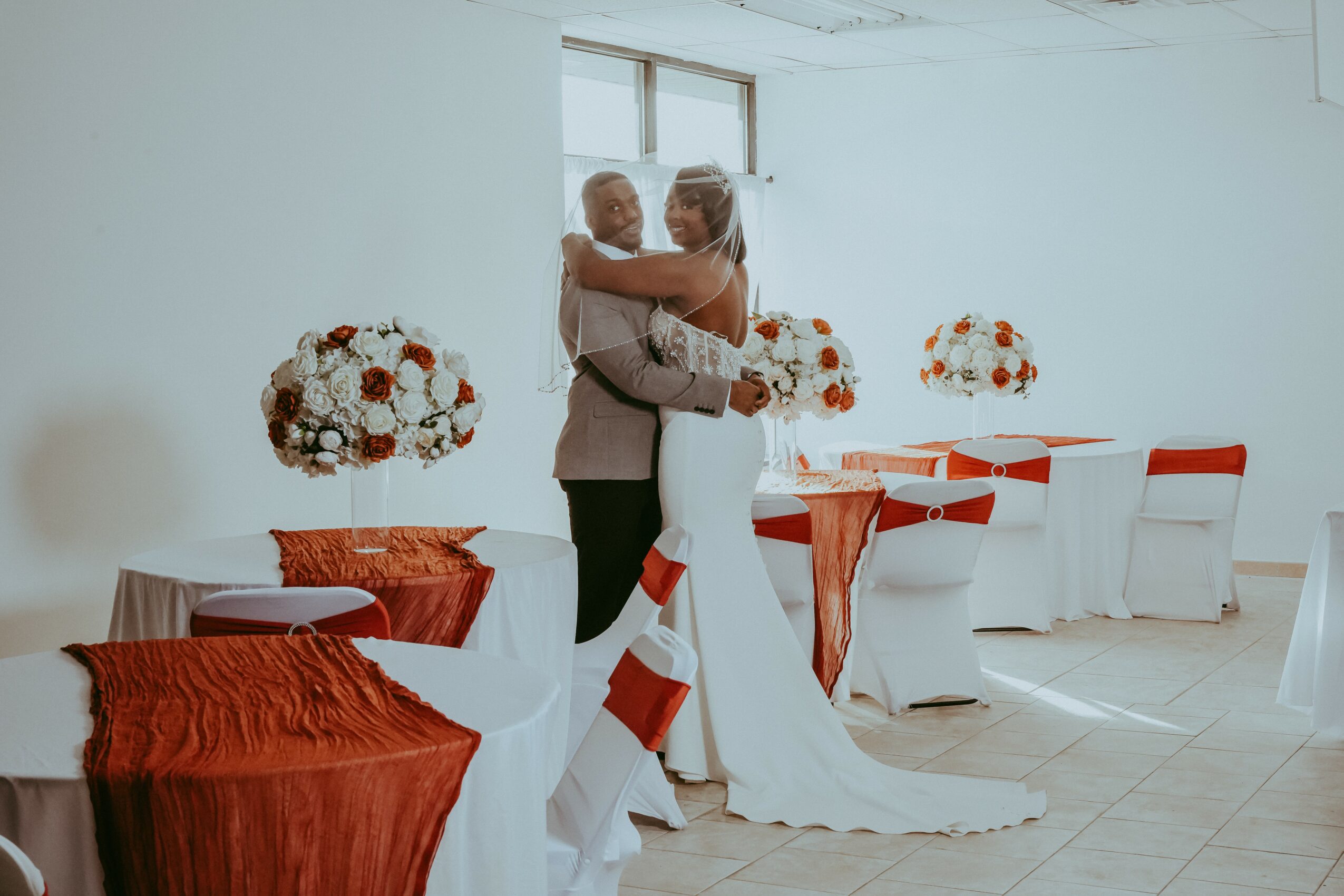 A couple dancing in a decorated event space with floral centerpieces and white chairs with red accents.