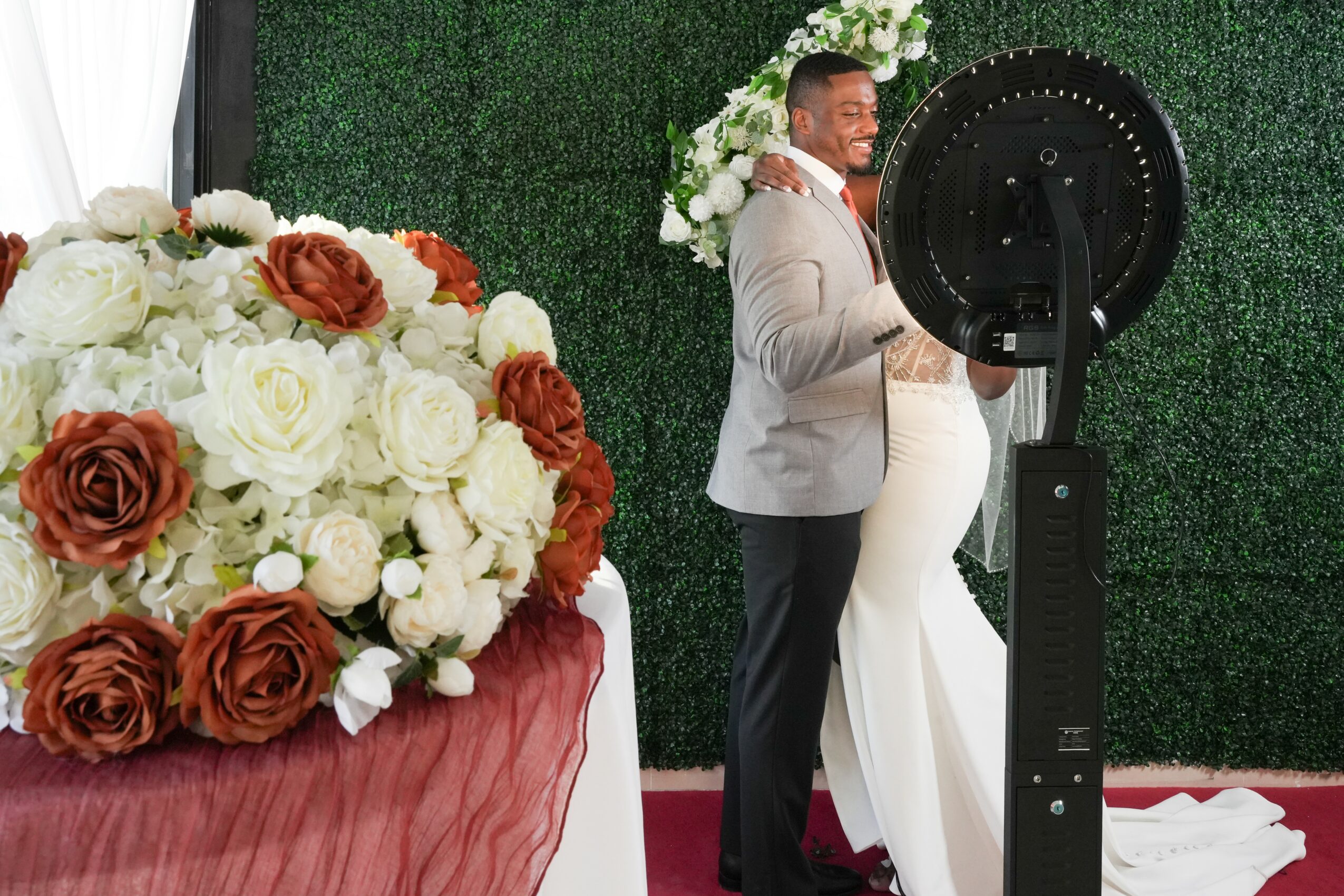 A man and woman in formal attire stand close together, smiling, with a large floral arrangement nearby, against a green backdrop.
