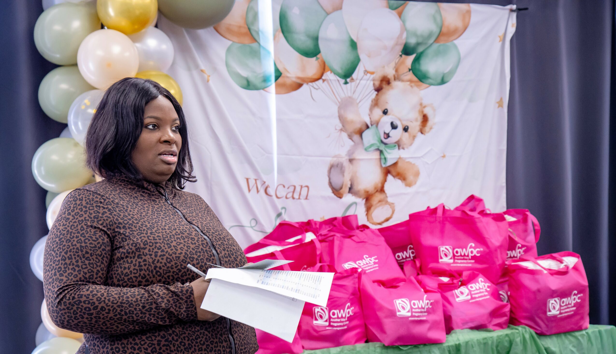 Woman holding papers in front of a decorated backdrop with balloons and pink gift bags.