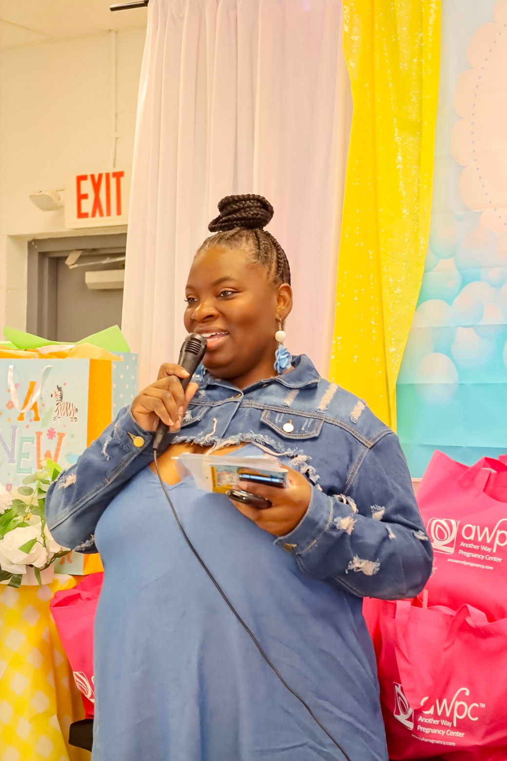Woman with braided hair in a denim jacket speaking into a microphone, holding a card, smiling, colorful background with pink and yellow decorations.