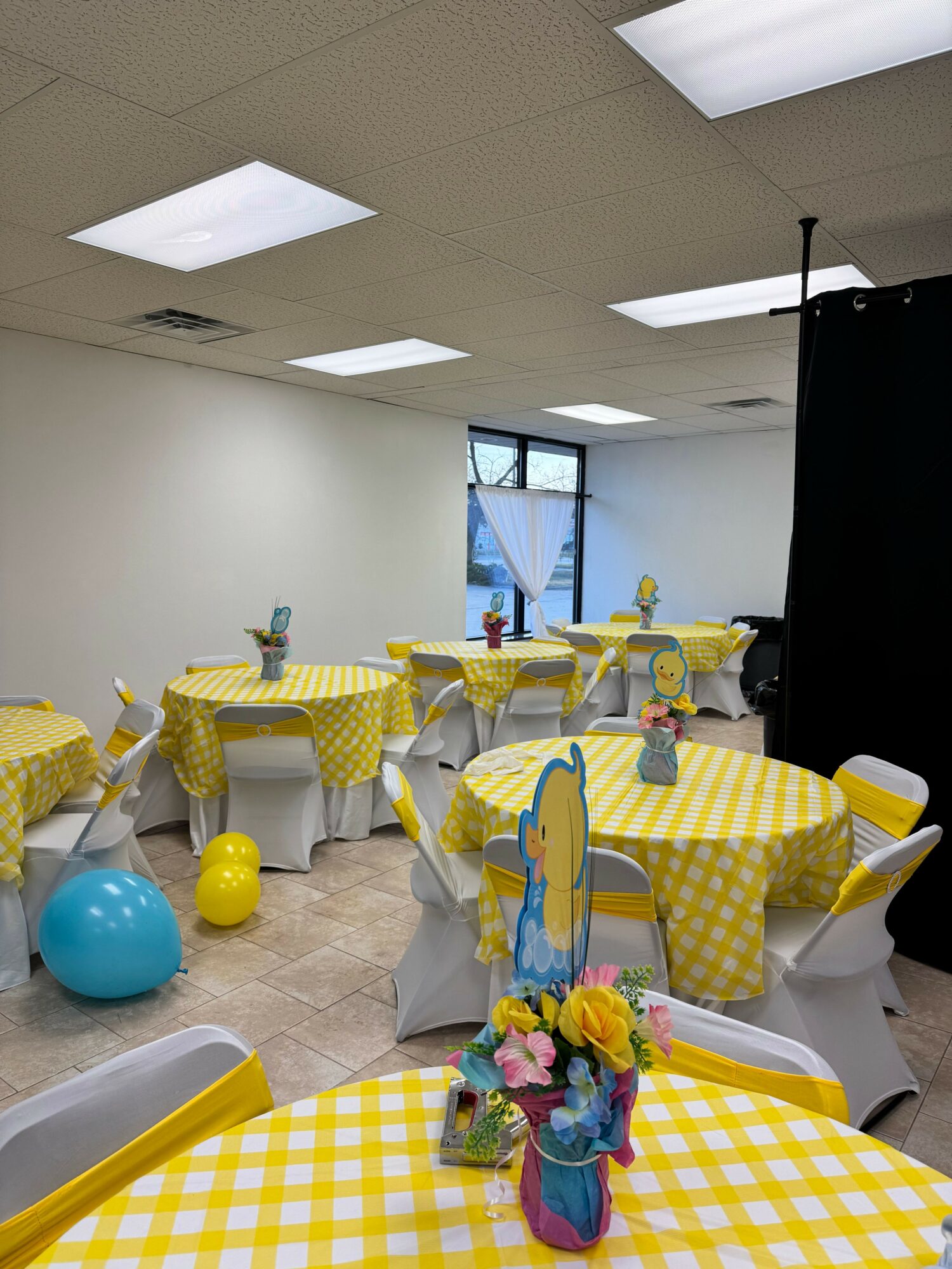 Party room with tables covered in yellow checkered tablecloths, decorated with balloons and floral centerpieces, set for celebration.