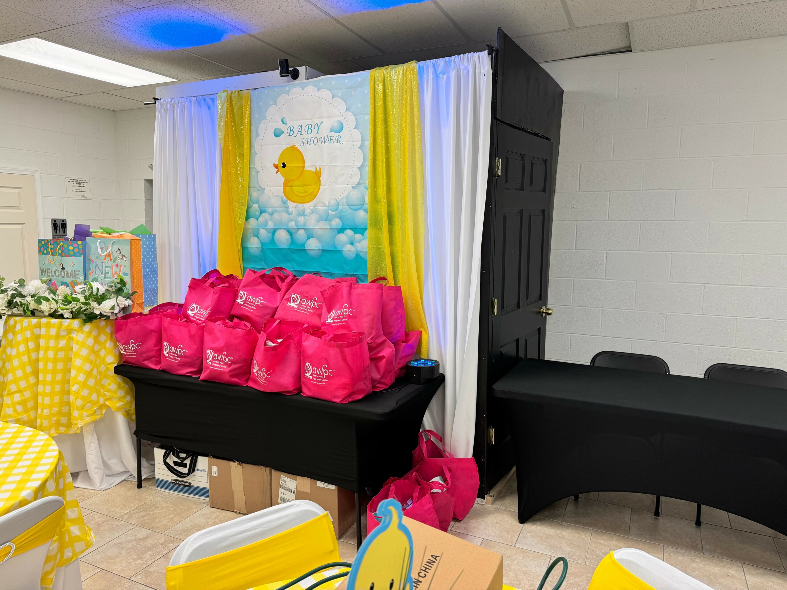 Baby shower setup with pink gift bags, yellow and white decorations, and a duck-themed backdrop.