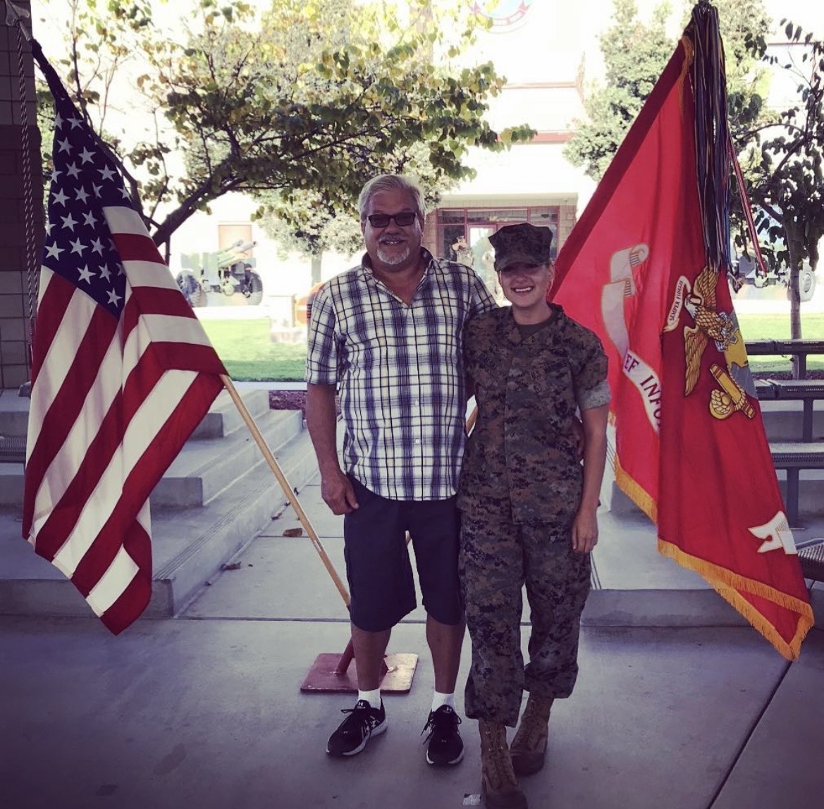 Two people standing outdoors, one in casual clothes and the other in military uniform, with American and Marine flags behind them.