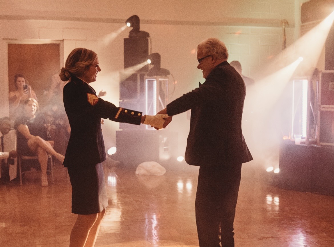 Two people dance and hold hands in a warmly lit room with a wooden floor, surrounded by onlookers.