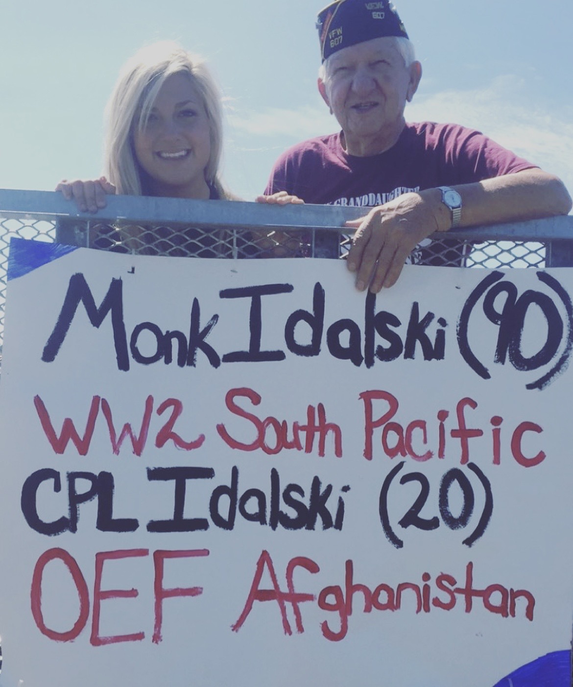 Two people holding a sign with handwritten text, smiling outdoors against a blue sky.