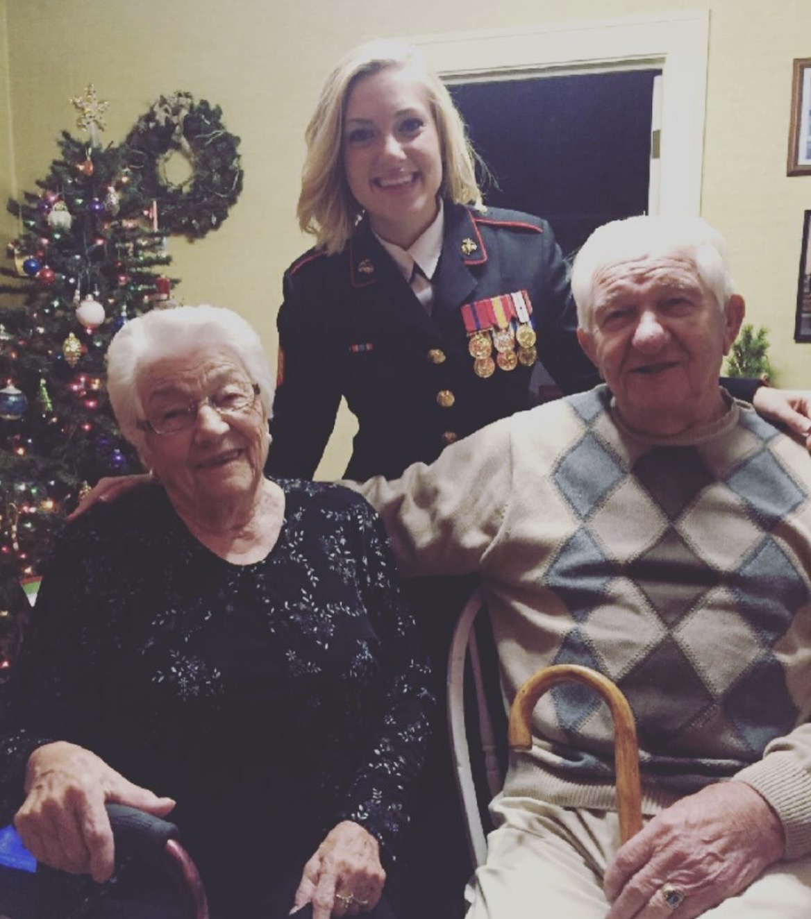 Three women, one in military uniform, smiling indoors near a decorated Christmas tree, with two elderly women seated and one standing behind.