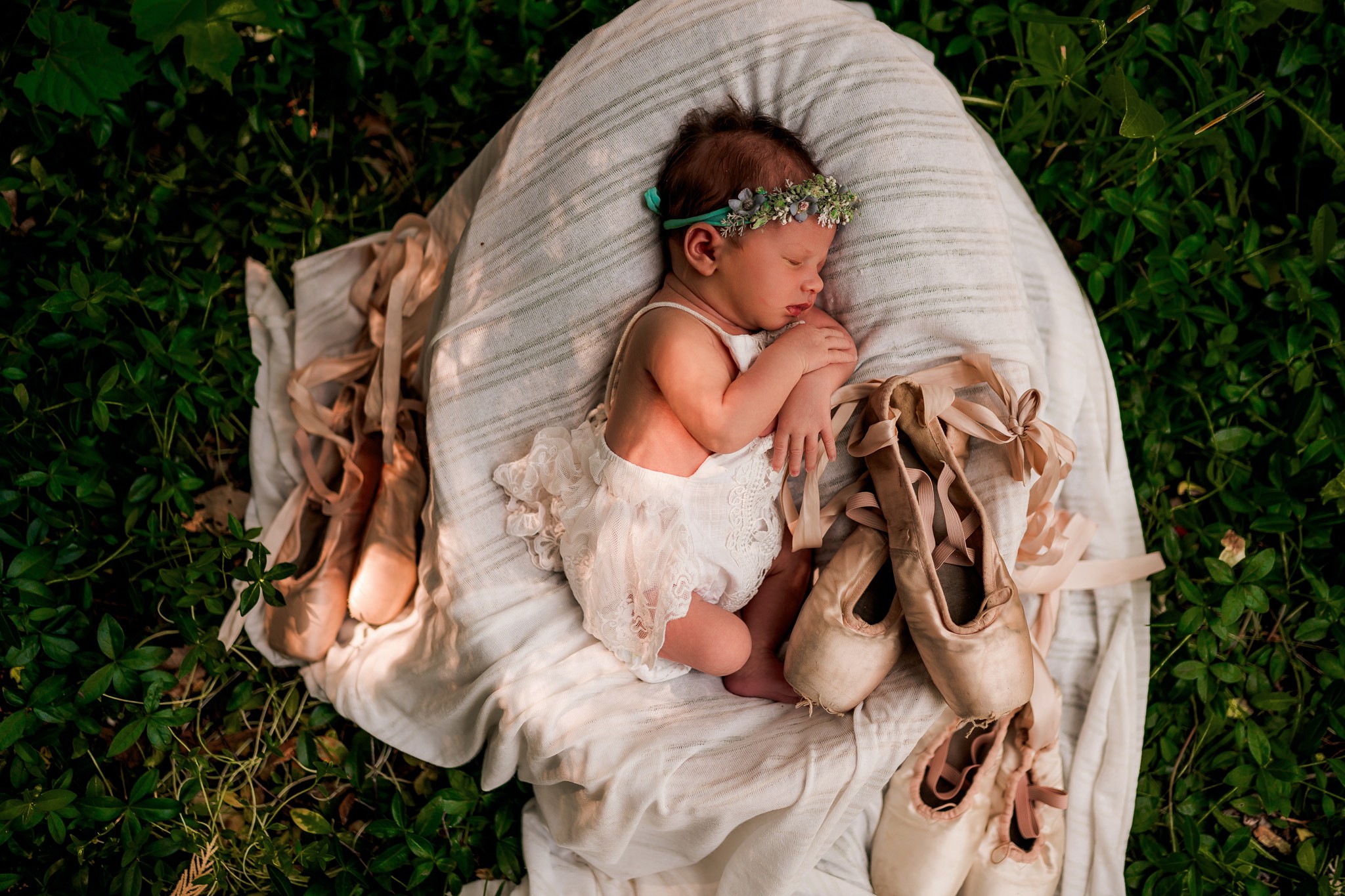 Baby girl lying on a white cushion outdoors, surrounded by green grass, with ballet shoes and a floral headband.