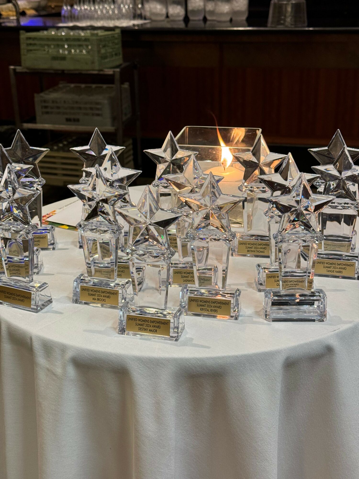 Table with star-shaped glass awards and small gold plaques, white tablecloth, background with wooden paneling.