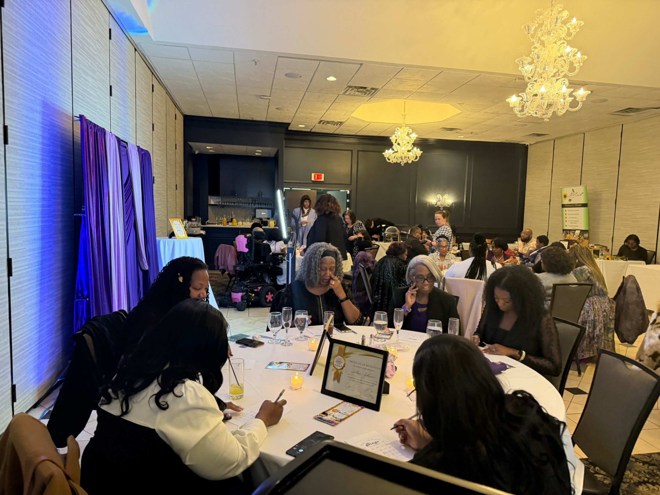 People seated at round tables in a banquet hall, some using phones, with chandeliers overhead and purple curtains on the wall.