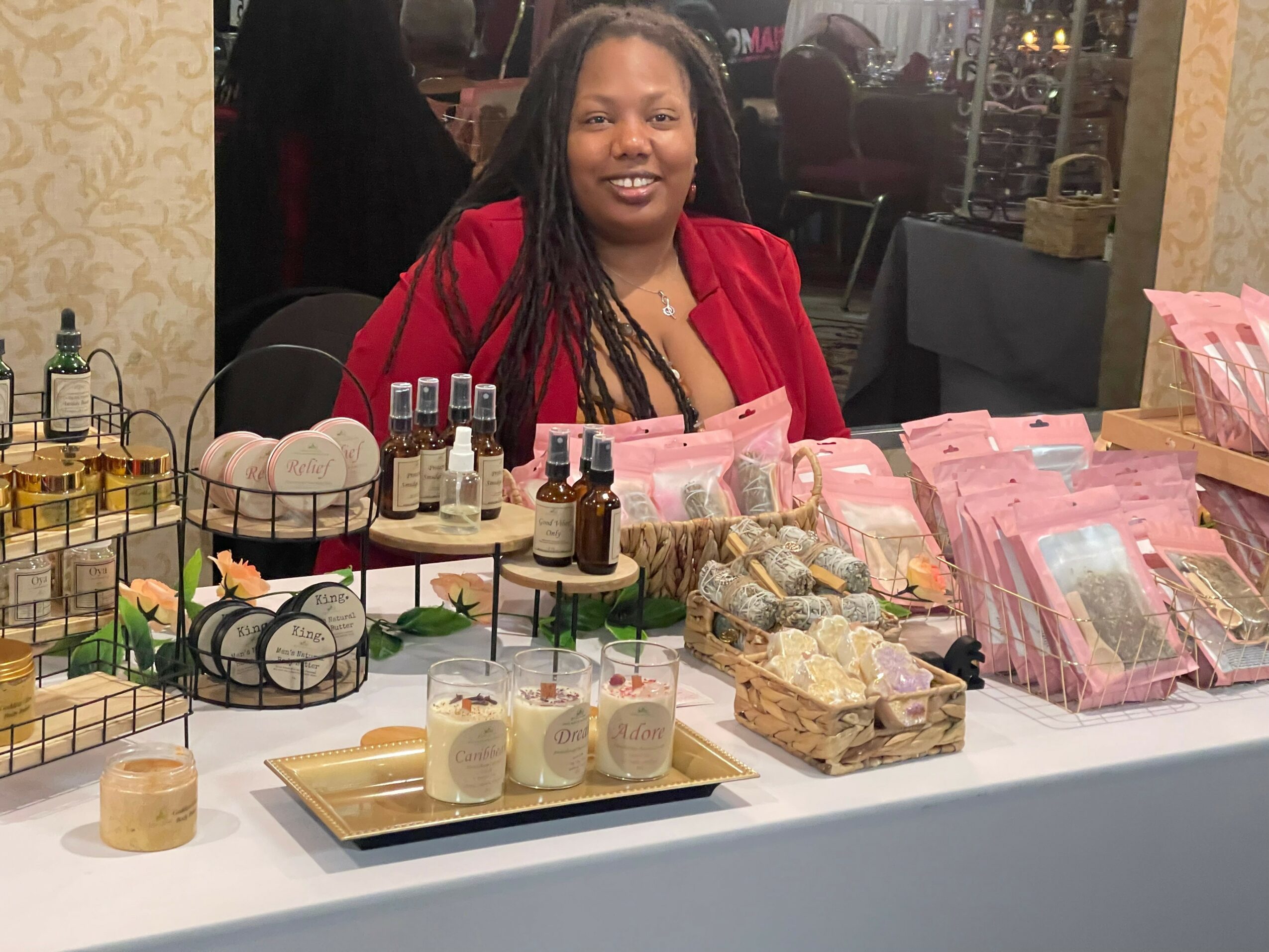 Smiling woman with long braids in red blazer behind a table with beauty products and packaged items.