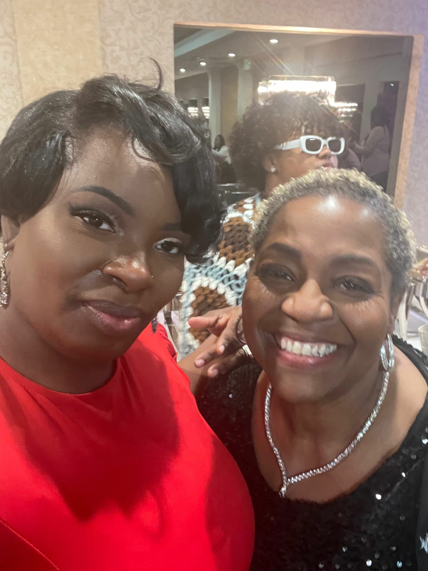 Two women smiling at a social event, one wearing a black dress with a necklace, the other in a red top.