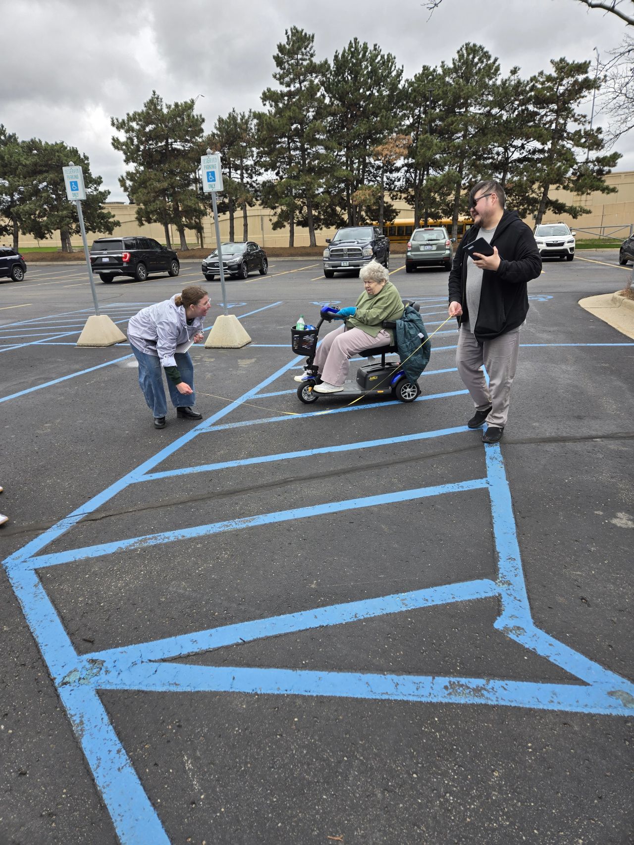 Three people in a parking lot: a person in a wheelchair, a child, and an adult, with parking spaces and trees in background.
