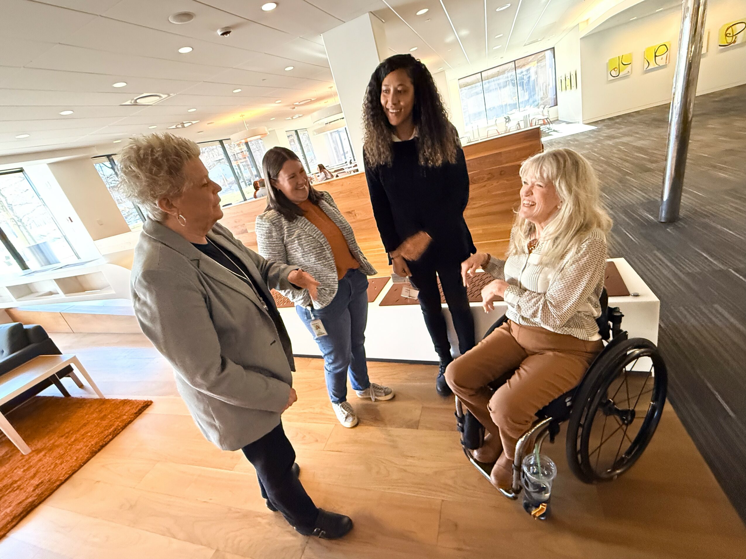 Four women are engaged in conversation in a modern indoor space, one woman in a wheelchair is part of the group, smiling.