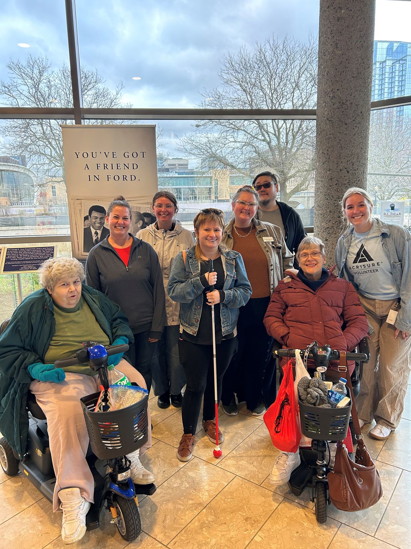 Group of nine people, some with mobility aids, inside a building with large windows and trees outside.