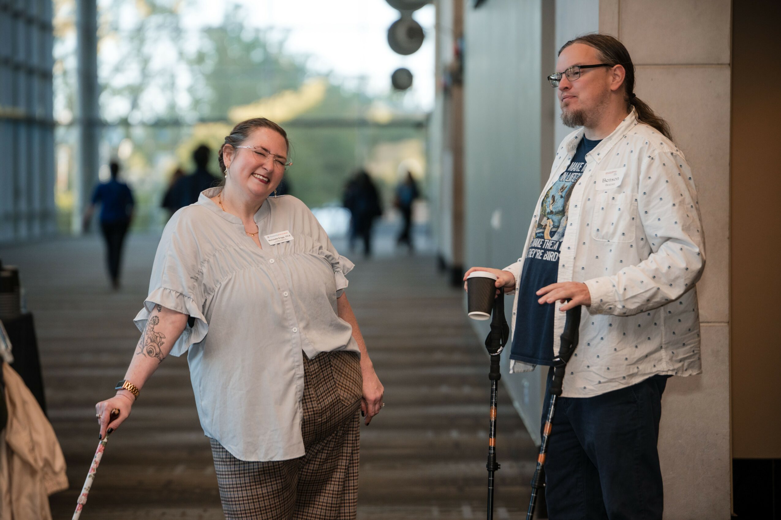 Two women standing and talking indoors, one smiling and holding a cane, the other with a cane and glasses.
