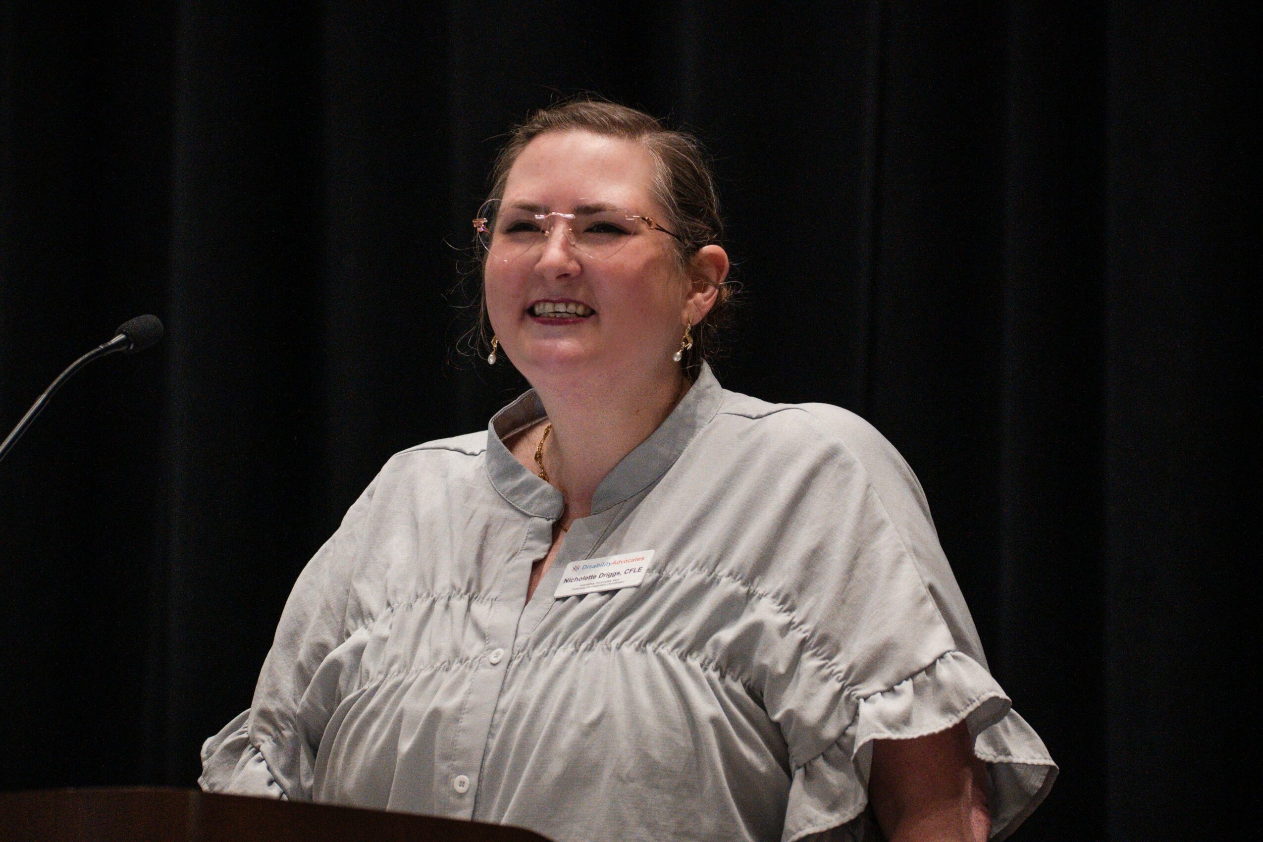 Smiling woman with glasses and earrings, wearing a light-colored blouse, standing in front of a black curtain.