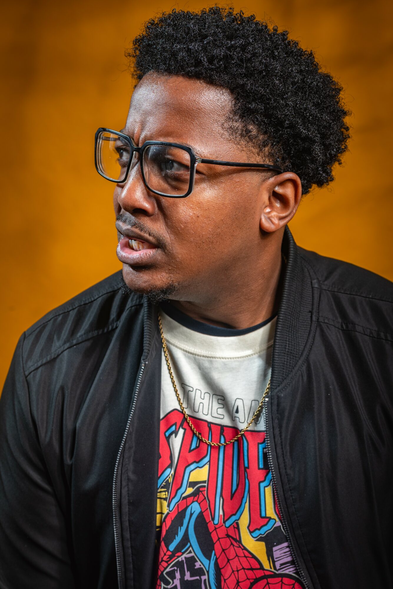 Man with glasses and curly hair wearing a black jacket and graphic t-shirt, looking to the side, yellow background.