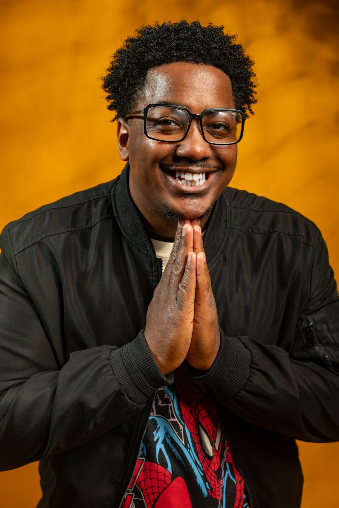 Smiling person with glasses and curly hair, hands pressed together, wearing a black jacket, yellow background.