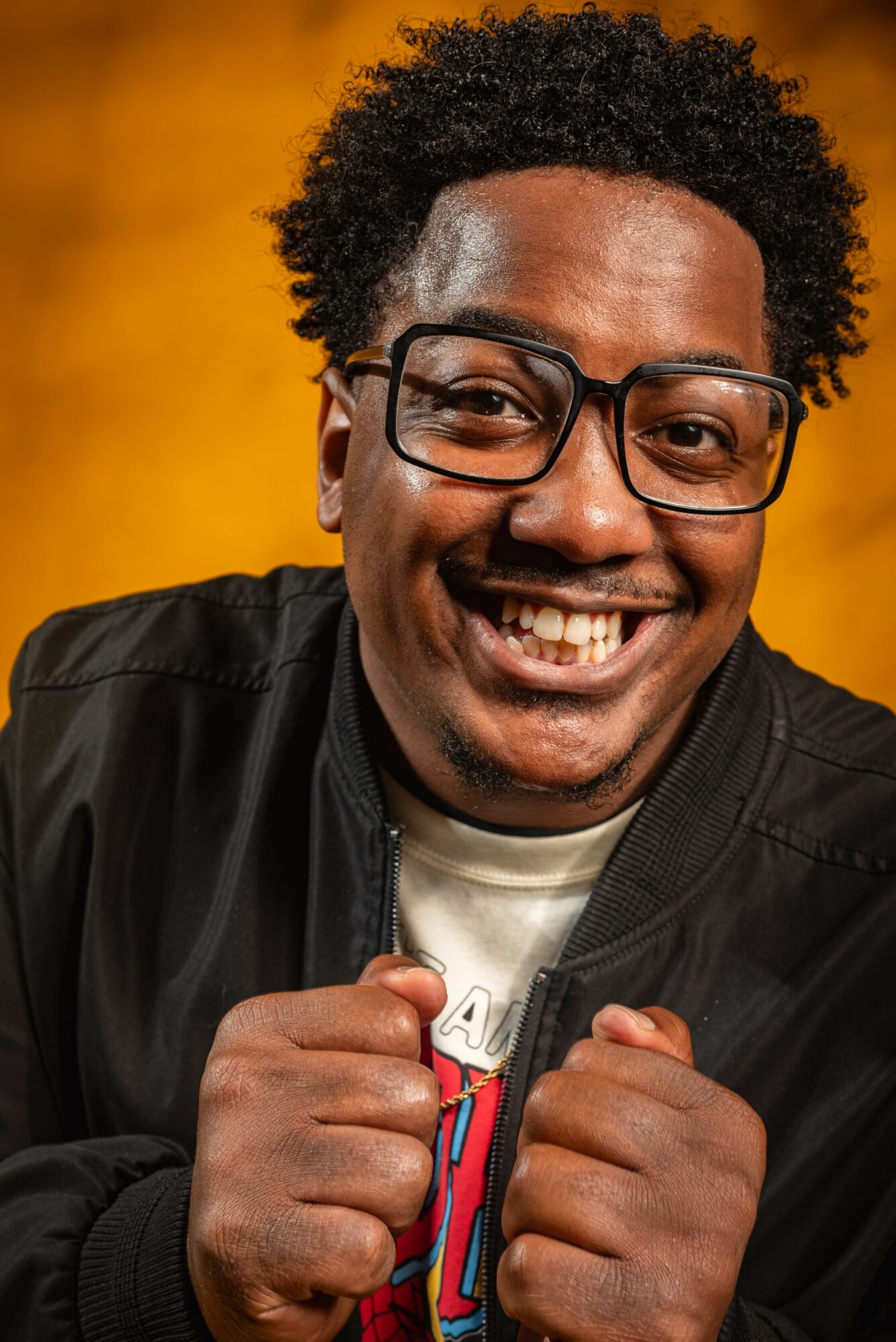 Smiling man with glasses and curly hair, wearing a black jacket, showing fists, against a yellow background.