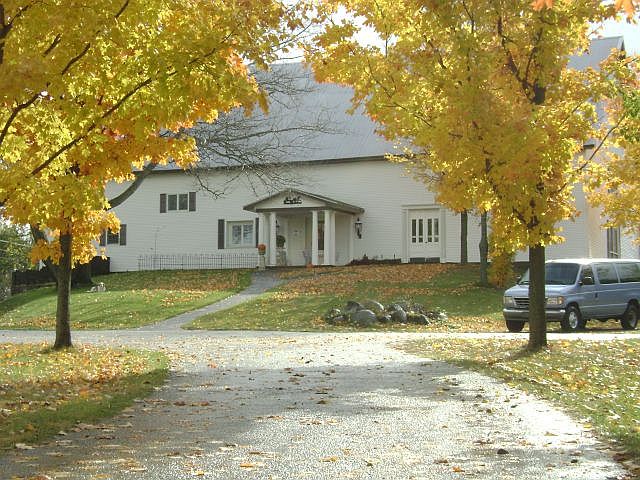 White house with front porch, surrounded by trees with yellow leaves, parked car on the right, and a pathway leading to the entrance.