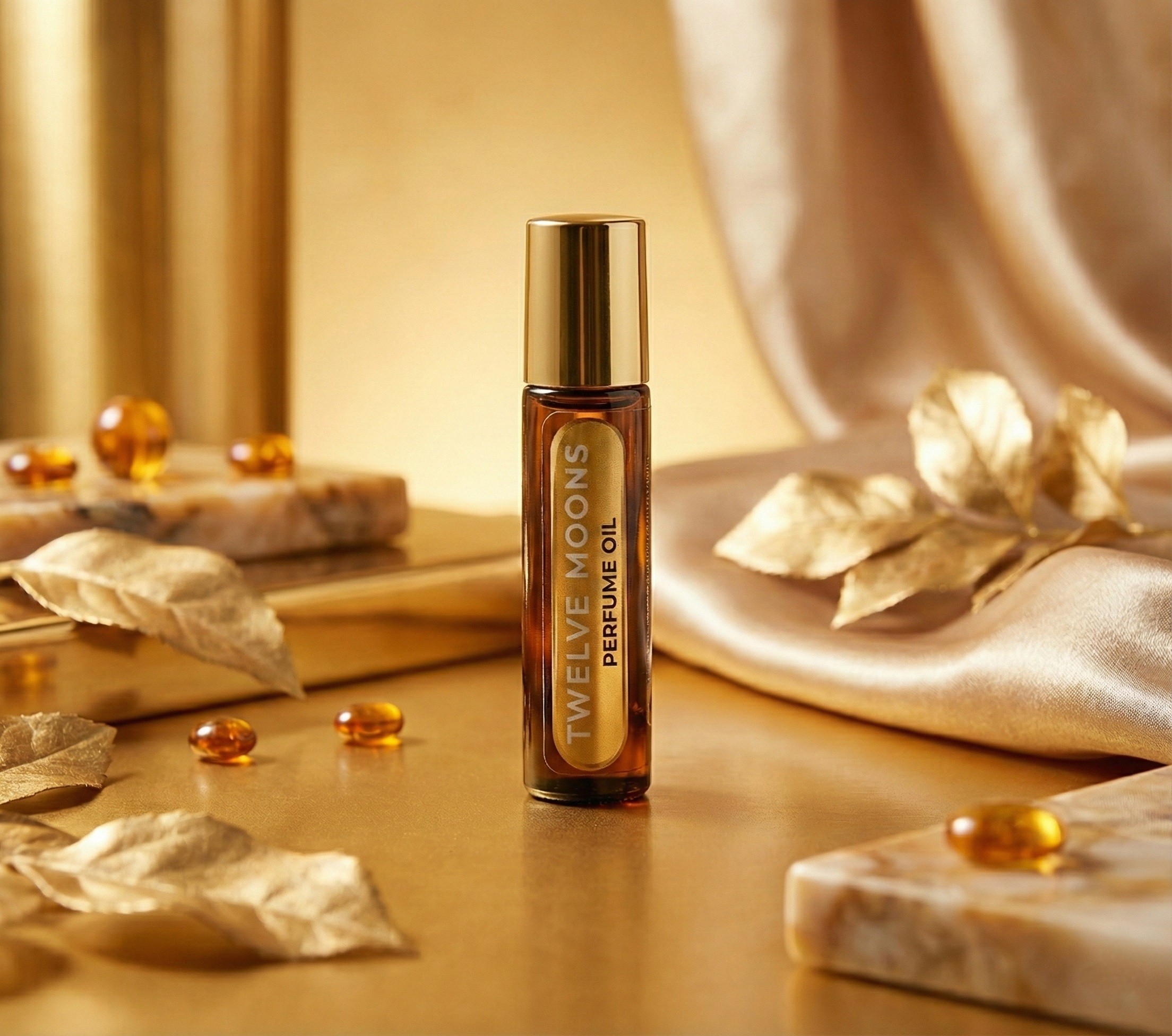 Small bottle of TWELVE MOONS perfume oil on a gold surface with decorative leaves and beads, warm background.