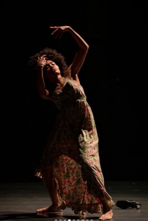 Person dancing on stage with one arm raised and head tilted, wearing a floral dress, dark background.