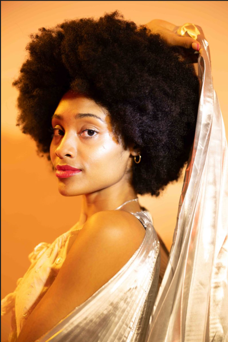Portrait of a woman with natural curly hair, wearing a shiny silver dress, against a warm orange background.