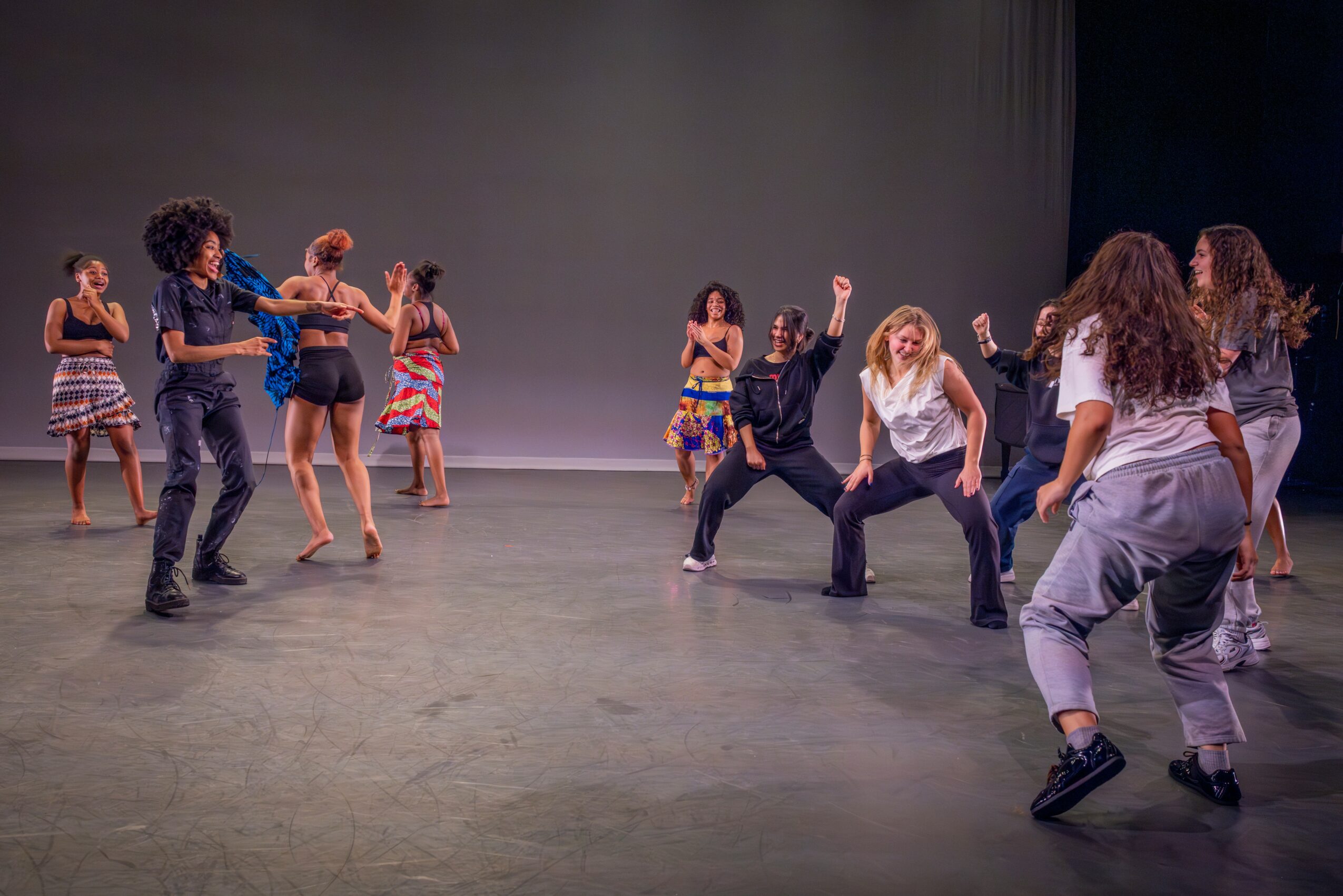Group of children and teenagers dancing on stage with a plain background, some in colorful outfits and others in casual clothes.