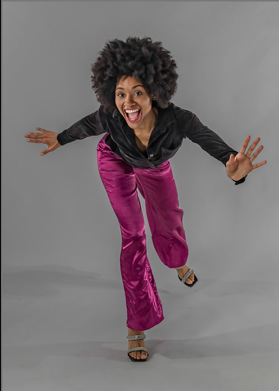 Woman with curly hair in black top and purple pants, smiling and reaching forward, standing on one foot against a gray background.