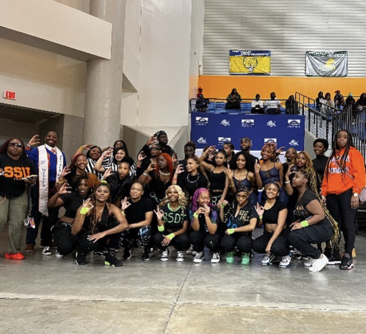 Group of people posing for a photo indoors, some making peace signs, with a blue backdrop and spectators in the background.