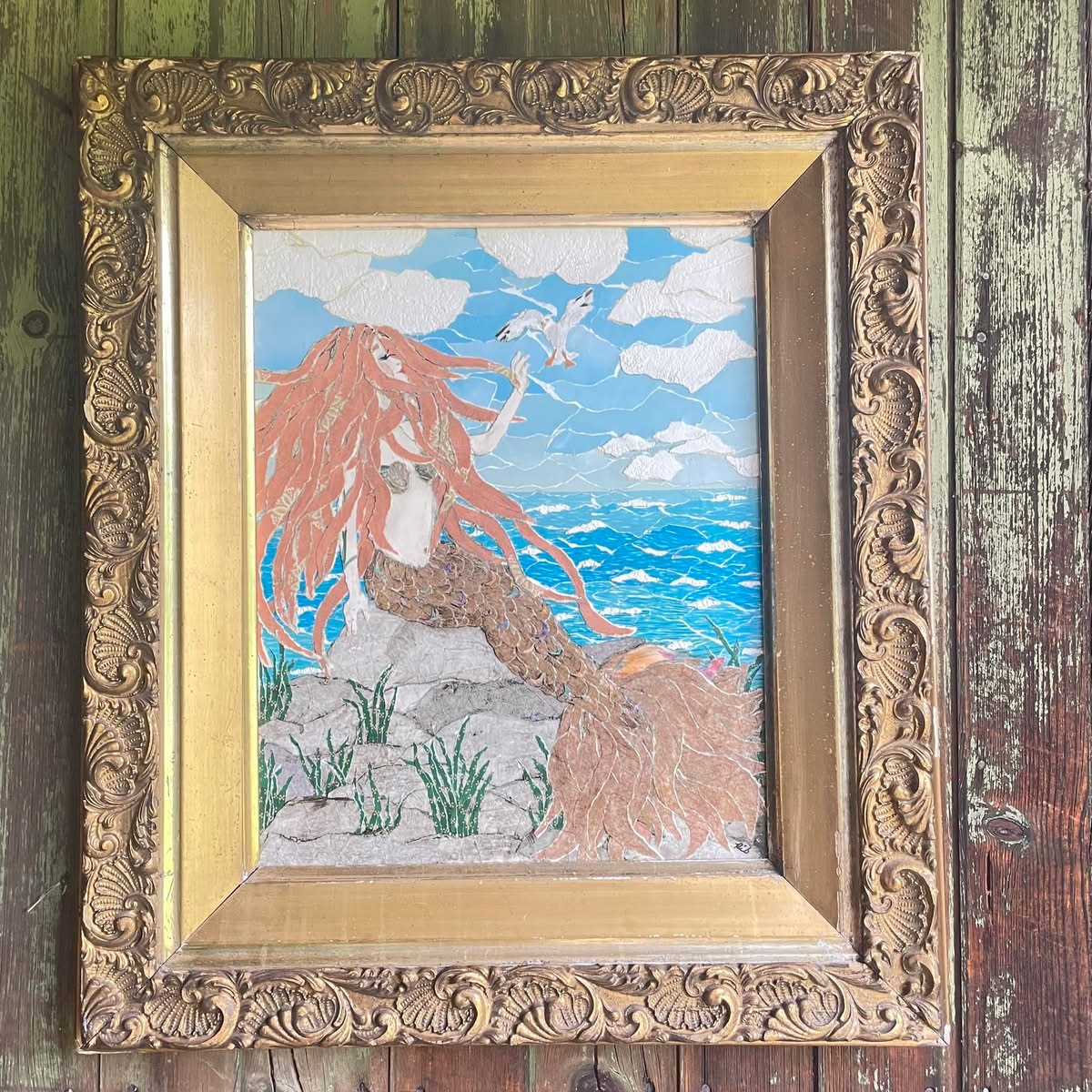 Painting of a mermaid with long red hair sitting on rocks by the sea, with clouds and blue sky in background.