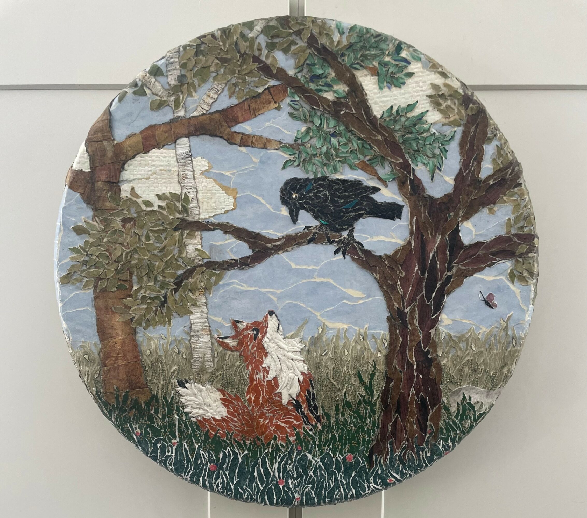 Circular embroidered scene with a fox lying on grass, a black bird on a tree branch, and a ship in the background.