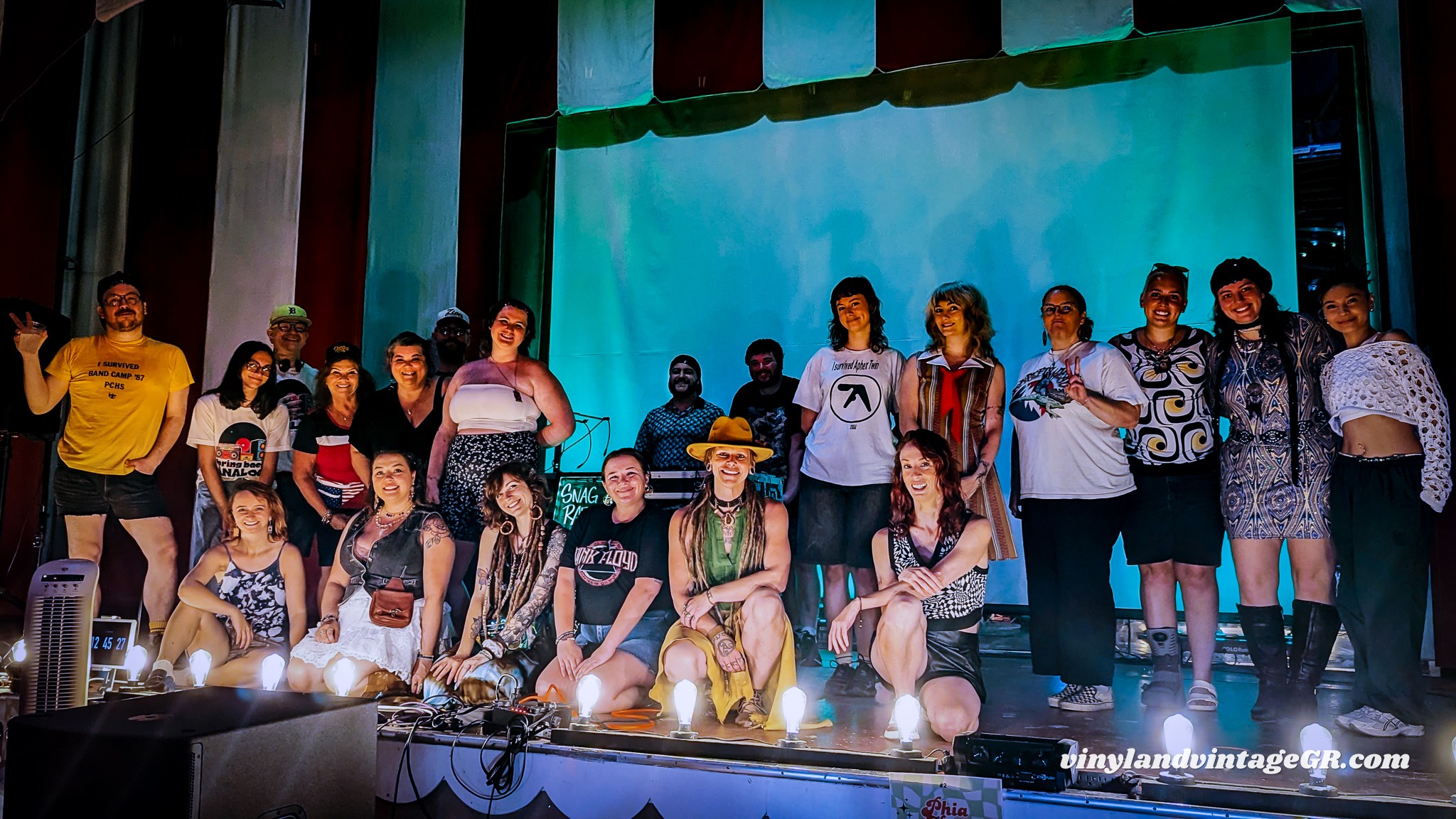 Group of people on stage, some sitting and some standing, with a large screen behind them, stage lighting visible.