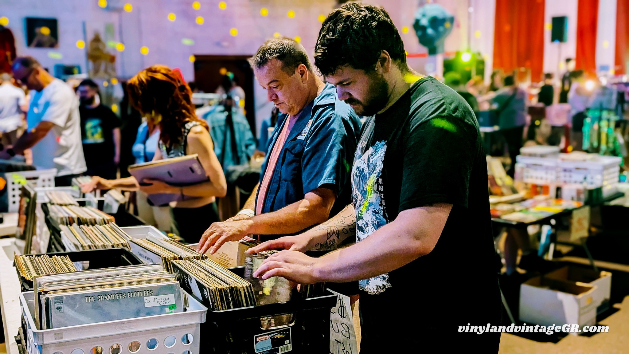 Two men browse vinyl records at a busy indoor market or store, surrounded by other shoppers and displays.