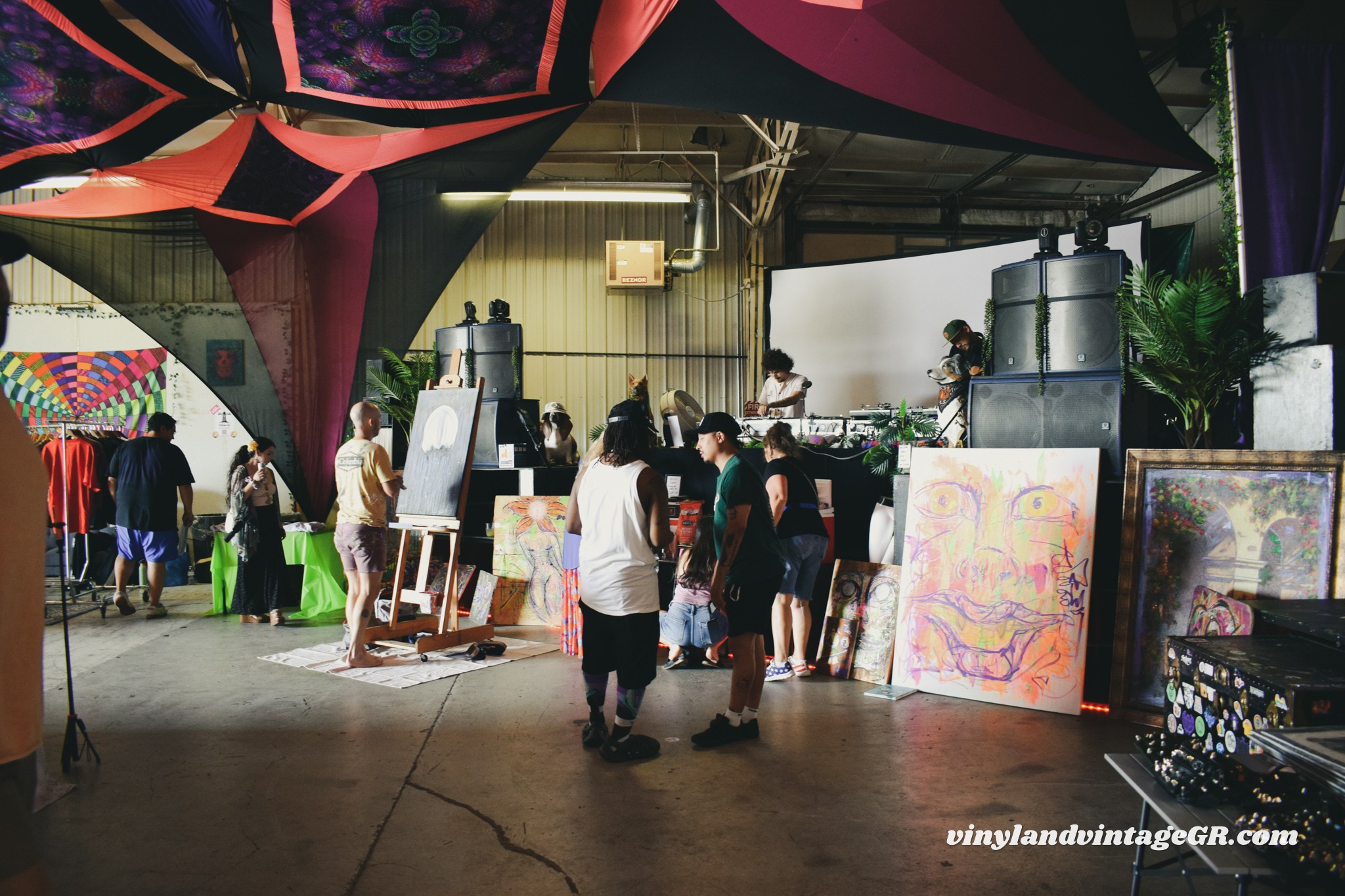 People gather at an indoor event with artwork and displays, under colorful fabric decorations hanging from the ceiling.