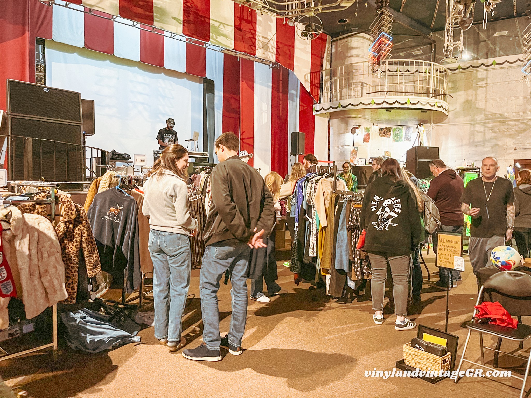People browsing clothing racks at an indoor market with red and white striped decorations and a stage in the background.