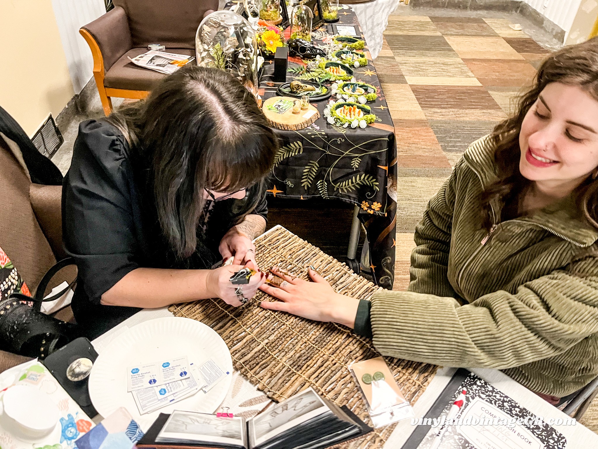 Two women sit at a table, one applying nail polish to the other's hand, with a spread of food and flowers in the background.