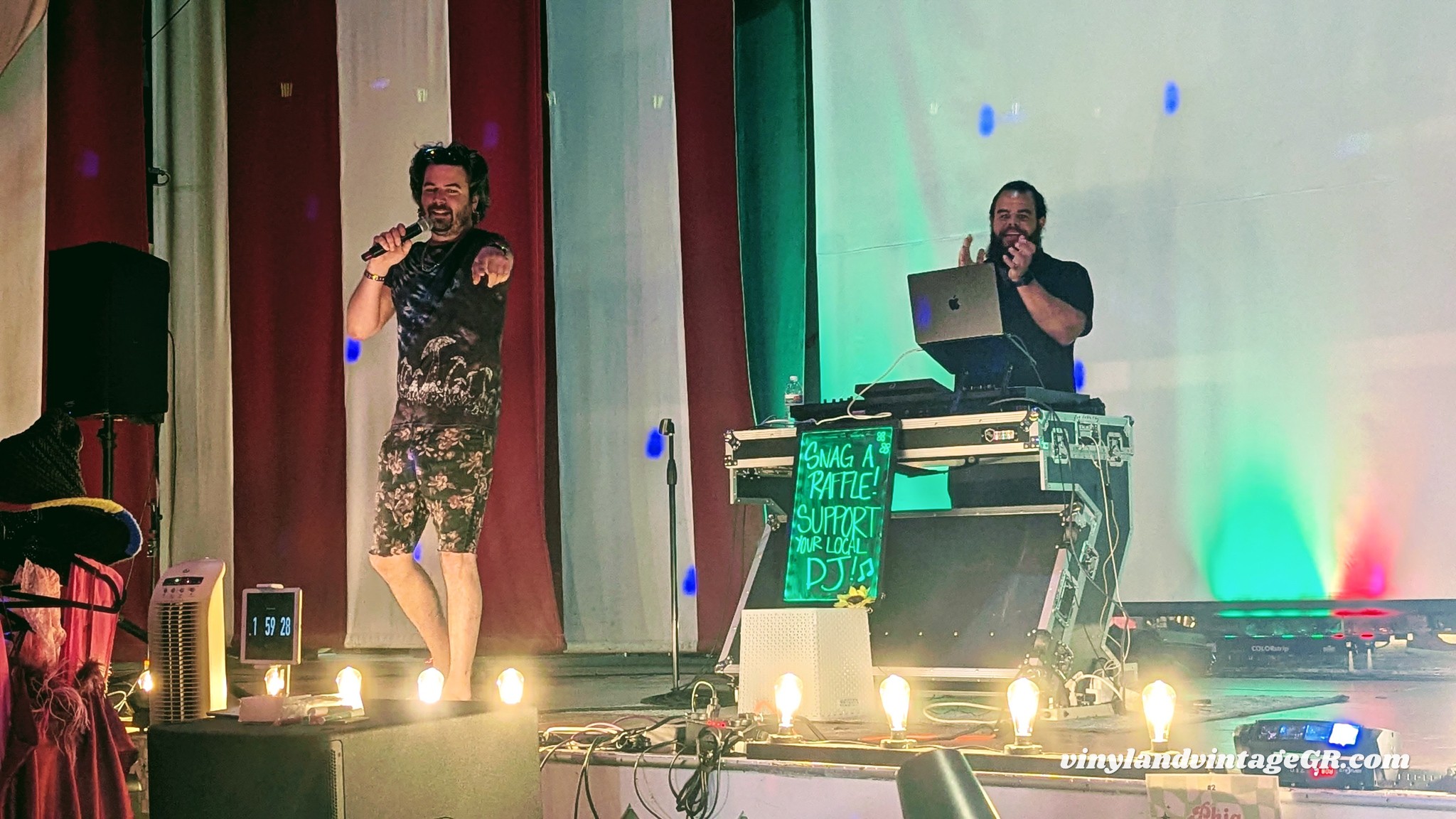 Two performers on stage, one singing with a microphone, the other DJing with a laptop, colorful lighting, and curtains in background.