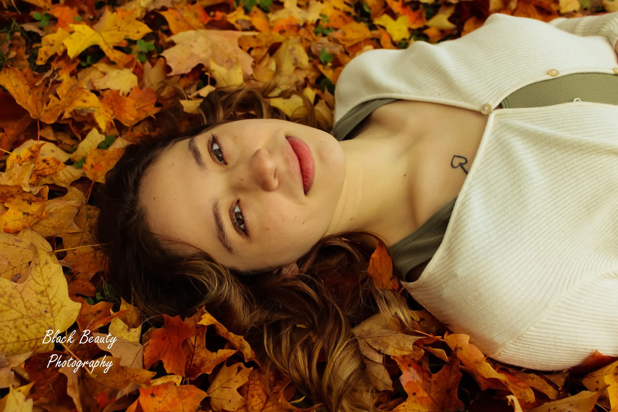 Woman lying on autumn leaves, looking at camera, with long hair and light-colored clothing.