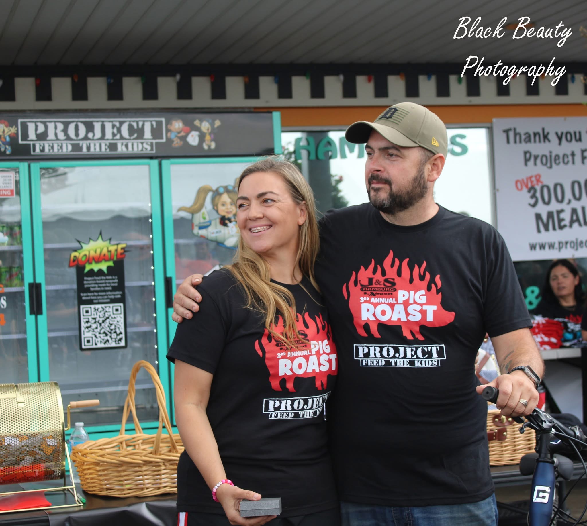 Two people wearing black shirts with a pig roast logo, smiling at an outdoor event, with banners and tables behind them.